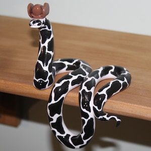May include: A handcrafted snake figurine with a black and white spotted pattern, wearing a small brown cowboy hat. The figurine is displayed on a wooden surface. The design is unique and playful, perfect for a quirky home decor item.