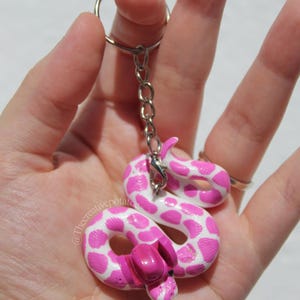 Könnte beinhalten: Handgefertigter Schlüsselanhänger mit einem rosa und weißen Schlangenanhänger. Die Schlange ist mit einem gepunkteten Muster bemalt und hat einen rosa Kopf. Befestigt an einer silbernen Kette und einem Schlüsselring.