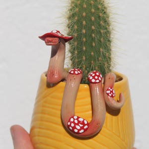 May include: A ceramic planter shaped like a snake wearing a red and white mushroom hat. The snake is curled around a cactus in the planter. The planter is yellow with a textured surface.