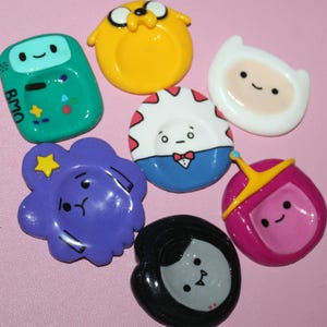Puede incluir: Colección de pines de personajes de Adventure Time de colores. Los pines presentan personajes como BMO, Jake, Finn, la Princesa Chicle y otros. Cada pin tiene un acabado brillante y está sobre un fondo rosa.