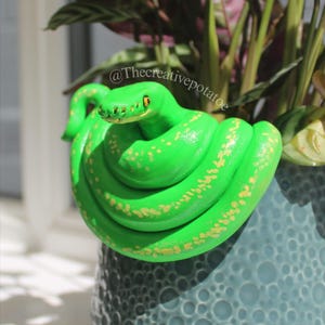 May include: A vibrant green snake sculpture with yellow spots is coiled around a blue ceramic pot. The pot has a textured surface and contains green and burgundy plants. The snake has yellow eyes. The image includes the text "@Thecreativepotatoe".