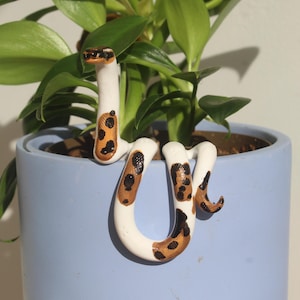 May include: A white snake figurine with brown and black leopard-like spots draped over the rim of a light blue cylindrical plant pot. The snake has a smooth finish and is positioned with its head raised upright against the green plant leaves.