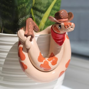 Könnte beinhalten: Handgefertigte Keramik-Schlangenskulptur, verziert mit einem Miniatur-Cowboyhut und einem roten Halstuch. Die Schlange ist cremefarben mit orangefarbenen Flecken bemalt und um einen weißen Topf gewickelt. Die Cowboy-Stiefel sind ebenfalls sichtbar.
