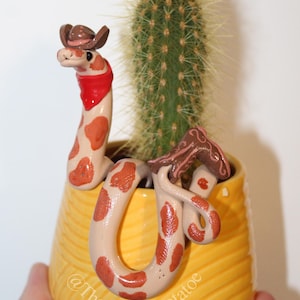 May include: A whimsical, handmade ceramic snake planter with a cowboy hat and red bandana, coiled around a cactus in a yellow pot. The snake has brown spots and is a unique decorative item.