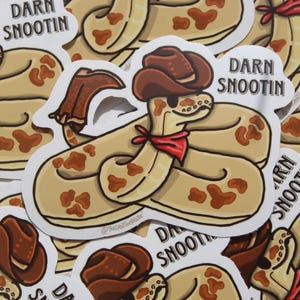 Könnte beinhalten: Eine Sammlung von Aufklebern mit einer Cartoon-Schlange, die einen Cowboyhut und ein rotes Halstuch trägt. Die Schlange ist hellbraun mit braunen Flecken. Der Text "Darn Snootin" ist auf weißem Hintergrund gedruckt.