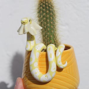 Könnte beinhalten: Ein gelber Keramikübertopf mit strukturiertem Diagonalmuster, der einen grünen Kaktus und eine gelb-weiße Schlangenfigur mit blumenförmigem Kopf enthält. Der Text @Thecreativepotatoe ist auf dem Körper der Schlange sichtbar.