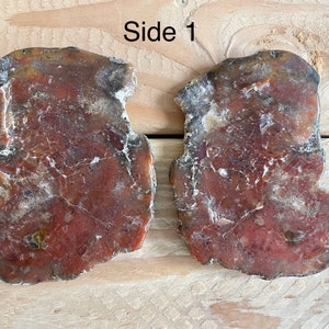 May include: Two irregularly shaped, red and white agate slices with a rough, natural edge. The slices are on a wooden surface. The text "Side 1" is visible above the slices.