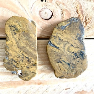 May include: Two irregularly shaped, flat pieces of polished stone with a swirling pattern of gold and black. The stone has a natural, organic look.