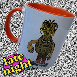 May include: White ceramic mug with an orange rim. The mug features a cartoon pineapple character with a green face and brown pants. The pineapple is wearing a brown hat and has a yellow and green pineapple top. The text "late night" is printed in pink and blue on the bottom of the mug.