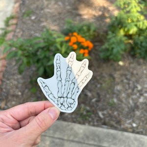 May include: A white sticker featuring a detailed illustration of a skeletal hand. The bones are depicted in black, creating a stark contrast against the white background. The sticker is held up against a blurred outdoor background.