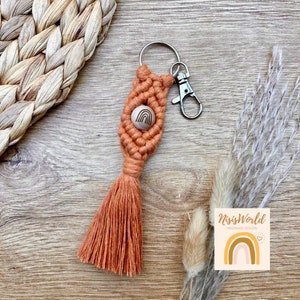 Könnte beinhalten: Ein orangefarbener Makramee-Schlüsselanhänger mit einer Holzperle, die ein Regenbogen-Design aufweist. Der Schlüsselanhänger hat einen silbernen Metallring und einen Karabiner.