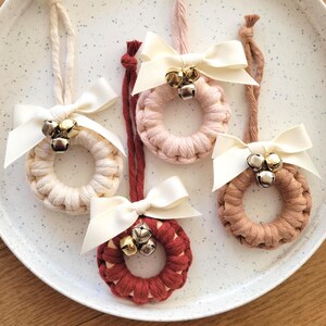 May include: Five macrame wreath ornaments in cream, pink, and burgundy, each with a cream-coloured bow and small gold bells. The ornaments are displayed on a speckled white plate, ideal for festive decoration.