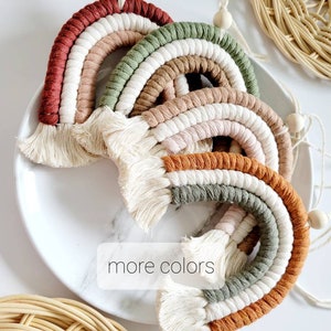 Puede incluir: Adornos de pared de arcoíris de macramé en varios colores, incluyendo óxido, verde y naranja. Cada arcoíris tiene un diseño en capas con flecos de borlas. La imagen incluye el texto "more colors".