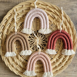 Puede incluir: Cinco adornos de arcoíris de macramé en tonos rosa, marrón y rojo, cada uno con flecos blancos y una cuenta de madera. Los arcoíris se exhiben sobre un salvamanteles de mimbre tejido.