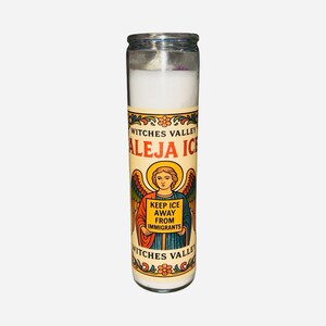 May include: A tall glass candle with a white wax interior. The label features the text "Witches Valley" and "Aleja Ice", along with an illustration of an angel holding a sign that reads "Keep Ice Away From Immigrants".