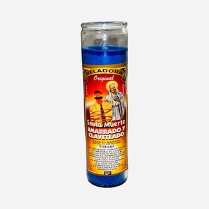 May include: A tall, cylindrical glass candle filled with deep blue wax. The label features a graphic of a person blindfolded with rope and a figure in a hooded robe, with text reading Veladora Original Santa Muerte Amarrado y Claveteado.
