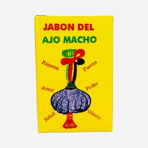 May include: A yellow box with the text "Jabon del Ajo Macho" in black. The box features a purple garlic with a red and green tassel on top. The words "riqueza", "fuerza", "amor", "poder", "salud", and "dinero" are written around the garlic.