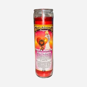 May include: A red glass candle with a label featuring the text "Veladora Original Santa Muerte Garrapata". The label includes an illustration of a heart, a figure, and additional text in Spanish. The candle is cylindrical.
