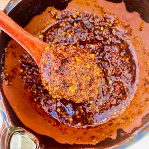 May include: A close-up of a terracotta bowl filled with a thick, reddish-brown sauce. The sauce is made with chili peppers, spices, and herbs. A wooden spoon is scooping out a portion of the sauce.