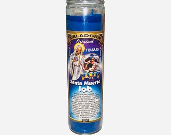 Puede incluir: Una vela de vidrio azul con una etiqueta que presenta el texto "VELADORA Original TRABAJO Santa Muerte Job". La etiqueta incluye una ilustración de una figura vestida y otras imágenes religiosas. La vela está llena de cera azul.