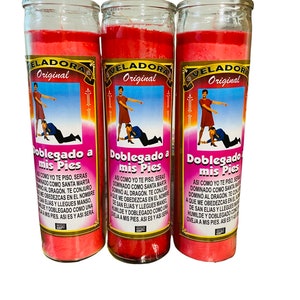 May include: Three red glass candle jars with a pink wax interior. Each jar has a label that reads "Veladora Original" and depicts a woman stepping on a man's back. The text on the label reads "Doblegado a mis Pies" and includes a prayer in Spanish.