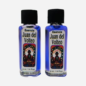 May include: Two small blue glass bottles with black caps. Each bottle contains a clear liquid and has a white label with the text "Esencia Juan del Volteo" and a colorful illustration of a man in a hat. The bottles are labeled with "8 ml. 1/4 Floz."