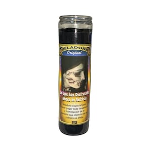 May include: A tall, cylindrical black candle in a glass jar. The label features the text "VELADORA Original" and a graphic of a face with a skull-like appearance. The text "Lo que has Disfrutado ahora lo Sufriras" is also visible.