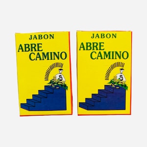 May include: Two rectangular soap boxes with bright yellow backgrounds and red borders. The text JABON ABRE CAMINO is printed in green. Each box features a graphic of a blue staircase leading to a white money bag with a dollar sign and gold coins.