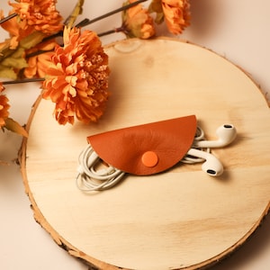 Könnte beinhalten: Ein brauner Leder-Kabelhalter mit einem orangefarbenen Knopf, der weiße Ohrhörer auf einer Holzoberfläche hält.