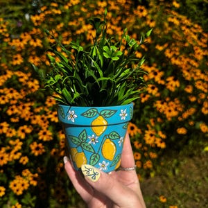 May include: A hand-painted, turquoise ceramic flower pot with a lemon and floral design, filled with green foliage. The background shows blurred yellow flowers and greenery. The pot is likely 10-15 cm in height.