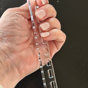 K&ouml;nnte beinhalten: Ein transparentes Kunststoff-Nagelkunst-Stempelwerkzeug mit mehreren Reihen kleiner, rechteckiger L&ouml;cher. Das Werkzeug ist so konzipiert, dass es Nagellack zum Stempeln von Designs auf N&auml;gel aufnehmen kann.