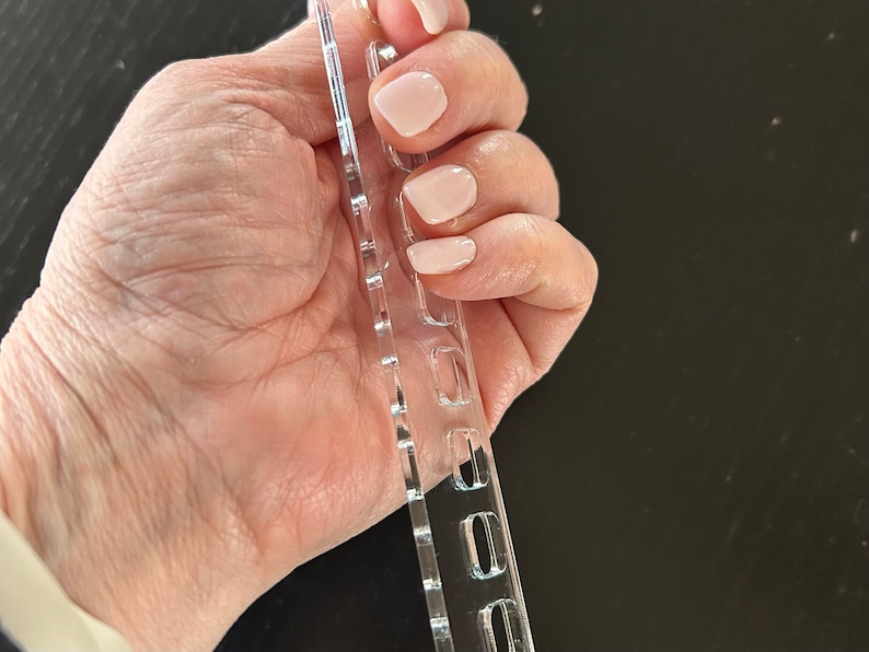 K&ouml;nnte beinhalten: Ein transparentes Kunststoff-Nagelkunst-Stempelwerkzeug mit mehreren Reihen kleiner, rechteckiger L&ouml;cher. Das Werkzeug ist so konzipiert, dass es Nagellack zum Stempeln von Designs auf N&auml;gel aufnehmen kann.