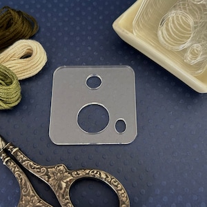 May include: A clear plastic template with three circular cutouts. The template is on a blue background with spools of thread.