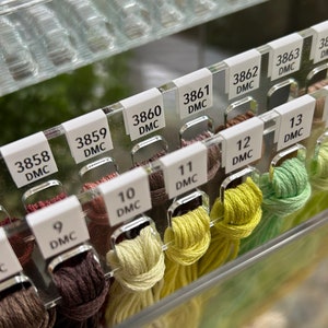 K&ouml;nnte beinhalten: Nahaufnahme eines durchsichtigen Plastikorganizers mit Reihen von DMC-Stickgarn. Das Garn ist nach Farbe und Nummer sortiert, mit Etiketten, die die DMC-Nummer und Farbe angeben. Das Garn ist in Braun-, Gr&uuml;n- und Gelbt&ouml;nen.