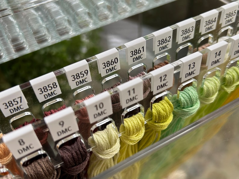 K&ouml;nnte beinhalten: Nahaufnahme eines durchsichtigen Plastikorganizers mit Reihen von DMC-Stickgarn. Das Garn ist nach Farbe und Nummer sortiert, mit Etiketten, die die DMC-Nummer und Farbe angeben. Das Garn ist in Braun-, Gr&uuml;n- und Gelbt&ouml;nen.