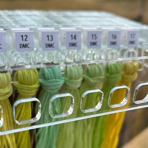 K&ouml;nnte beinhalten: Ein transparenter Plastik-Organizer mit F&auml;chern zur Aufbewahrung von Stickgarn. Die F&auml;cher sind mit den DMC-Nummern 11 bis 17 beschriftet. Das Garn ist um die F&auml;cher gewickelt und in Gr&uuml;n- und Gelbt&ouml;nen gehalten.