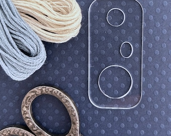 May include: A clear plastic template with three circles of different sizes. The template is on a dark blue background with a pair of silver scissors and two spools of embroidery floss.