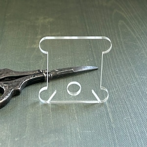 May include: A clear plastic spool holder for thread or yarn. The holder has a rounded top and a circular opening in the center.