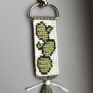 May include: A green, white, and beige woven keychain with a silver key ring and a green tassel. The keychain features a woven design of three green circles.