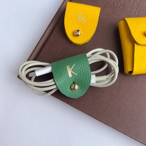 May include: A green leather cord organizer with a gold-toned snap button and the letter K embossed in gold, securing a white charging cable. Beside it are two yellow leather accessories, one featuring a gold snap and the letter K in gold.