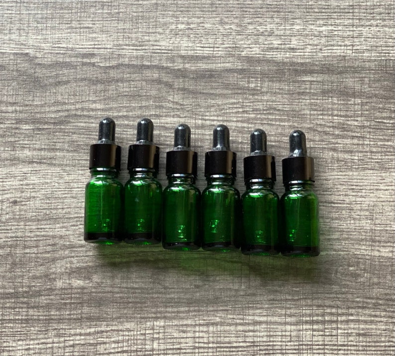 May include: Six small green glass bottles with black dropper tops. The bottles are lined up in a row on a wood surface.