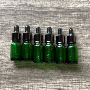 May include: Six small green glass bottles with black dropper tops. The bottles are lined up in a row on a wood surface.