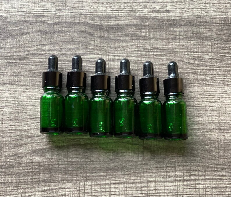May include: Six small green glass bottles with black dropper tops. The bottles are lined up in a row on a wood surface.