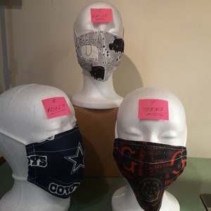 May include: Three face masks on white mannequin heads, labeled with pink sticky notes: "Adult", "Teens", and "Child". The masks are made of fabric with different patterns: a black and white animal print, a blue and white Dallas Cowboys pattern, and a black and red pattern with a circular design.