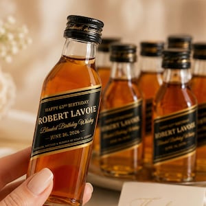 May include: A small glass bottle filled with amber liquid, featuring a black label with gold text that reads Happy 63rd Birthday, Robert Lavoie, Blended Birthday Whiskey, June 26, 2026, Distilled, Bottled & Blended by Kelly & Andrew.