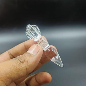 Op de afbeelding: Een heldere kristallen vajra, een ritueel object dat wordt gebruikt in het boeddhisme en hindoeïsme, met een puntige punt en een decoratieve bovenkant. De vajra wordt in een hand gehouden.