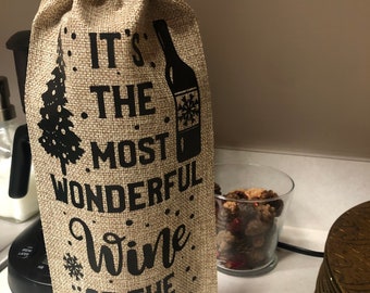 以下が含まれることがあります： 「It's the most wonderful wine of the year.」と黒文字で書かれた茶色のジュート製のワインバッグ。バッグは巾着式で、雪の結晶のデザインが施されています。