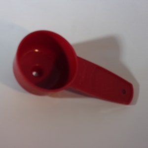 May include: A red plastic measuring scoop with a handle. The scoop has a small hole in the bottom.