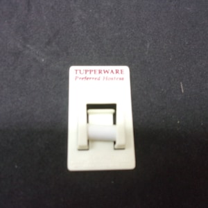 Puede incluir: Etiqueta blanca de plástico Tupperware Preferred Hostess con un logotipo rojo y un pequeño rodillo blanco de plástico.