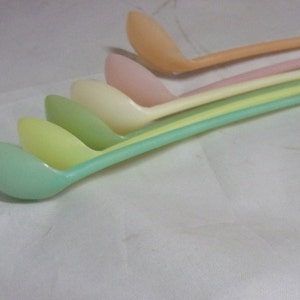 Puede incluir: Cinco cucharones de plástico de color pastel, dispuestos en un orden de arcoíris del naranja al verde, con los mangos superpuestos.